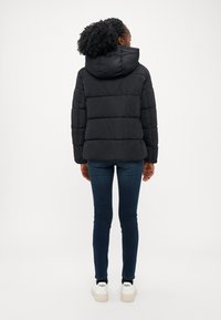 Svart polstret jakke med hette, preget av horisontal quilting, en avslappet passform og en jevn tekstur, kombinert med mørke jeans og hvite joggesko.