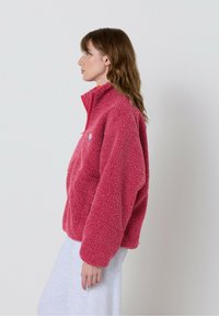 Flis pulover v živo roza barvi z visokim ovratnikom, zadrgo in sproščenim krojem. Teksturirana površina z majhno logotipno nalepko na prsih.