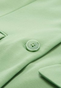Stof de un verde deschis, cu o textură netedă, prezentând un nasture rotund, asortat, cu patru găuri, situat lângă o buzunar cu clapă.