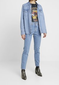 Camisa de mezclilla azul clara sobre una camiseta gráfica negra, combinada con pantalones vaqueros rectos de mezclilla azul clara y botas de tobillo negras con estampado de serpiente.