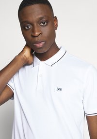 Giovane uomo che indossa una polo bianca Lee con rifiniture nere su colletto e maniche, in posa con una mano dietro al collo.
