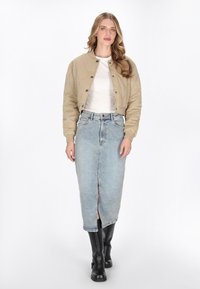 Kvinde iført beige quiltet jakke, hvid top, lyseblå denim midi-nederdel med forbidte slids og sorte knæhøje støvler, stående foran en hvid baggrund.