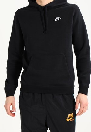 Osoba ubrana w czarną bluzę z kapturem Nike z białym logo oraz czarne spodnie dresowe Nike z żółtym logo, stojąca z rękami swobodnie opuszczonymi wzdłuż boków.