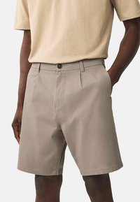 Mann in beigen, maßgeschneiderten Shorts mit Seitentaschen, der ein hellbeiges, kurzärmliges Hemd trägt, mit einer Hand in der Tasche steht.