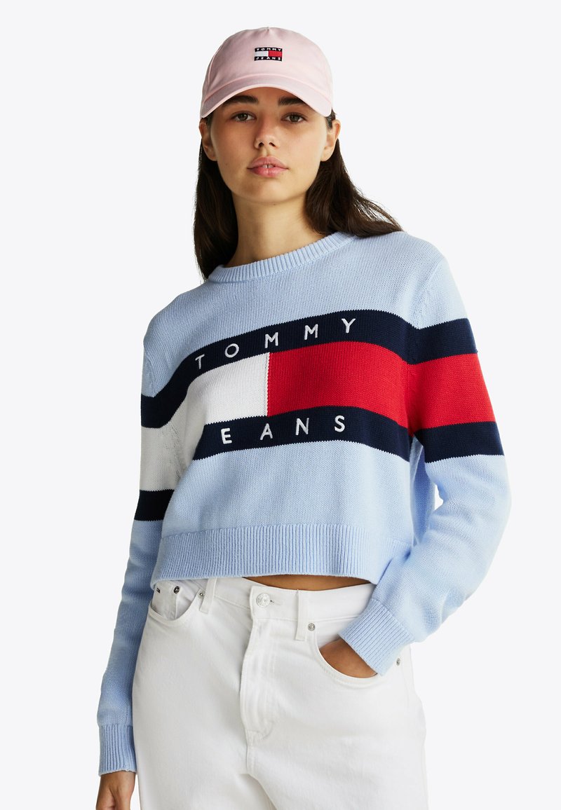 Lichtblauwe gebreide trui met marineblauwe, rode en witte strepen. Voorzien van het "TOMMY JEANS" logo over de borst. Gecombineerd met witte, hooggetailleerde broek.