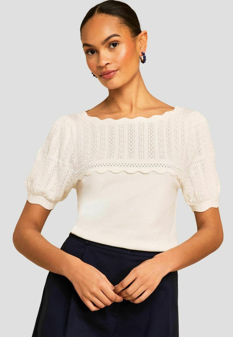 Naine kannab valget kootud õhulise varrukaga pluusi pitsdetaili ja lainelise äärega, tumeda kõrge vöökohaga seeliku seltsis, neutraalsel taustal.