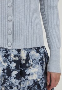 Cardigan en tricot côtelé bleu clair avec cinq boutons ronds, associé à une jupe à motifs floraux marine avec des nuances de bleu et de blanc.