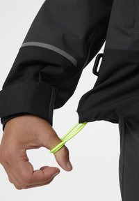 Černý rukáv bundy s reflexním šedým pruhem; neonově zelená elastická šňůra ovázaná kolem palce pro nastavení; voděodolná textilní struktura.
