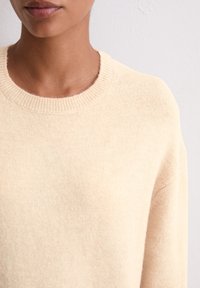 Pull en tricot beige avec un col rond, texture douce et col côtelé. Le tissu semble léger et légèrement duveteux.
