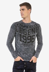 Langärmliges graues T-Shirt mit einem verblassten, strukturierten Design und schwarzem Drucktext "LEGEND 1996" sowie Akzenten. Kombiniert mit dunkler Hose.
