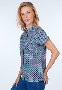 Short-sleeved blouse in vibrant blue, red, and green wave pattern. Features a collared neckline and front button closure. Smooth fabric texture.