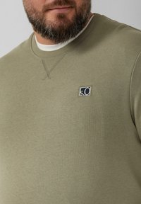 Felpa verde oliva con colletto a girocollo a costine e un piccolo patch con logo blu scuro sul petto. Tessuto morbido e testurizzato con dettagli di cucitura.