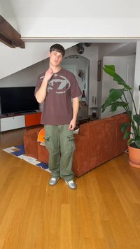 Jonge man in casual kleding staand in een woonkamer met een oranje bank, een groene kamerplant, een houten vloer en een televisie op een kast.