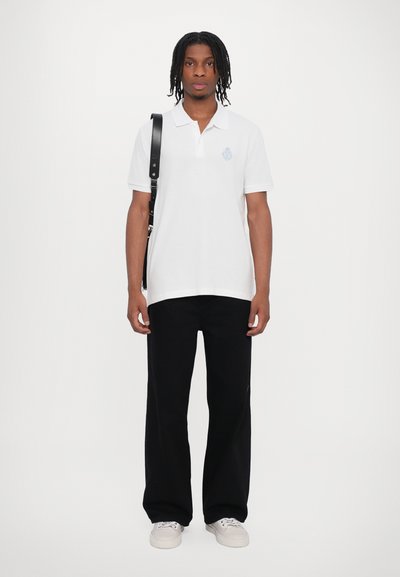 Camisa polo de algodón blanca con cuello y logotipo bordado en azul claro, combinada con pantalones de pierna ancha negros y zapatillas blancas.