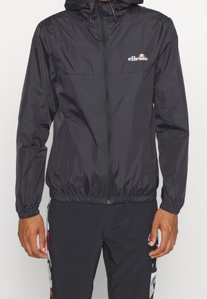 Uomo che indossa una giacca con cappuccio nera con zip e logo ellesse sul petto e pantaloni neri con strisce laterali colorate.