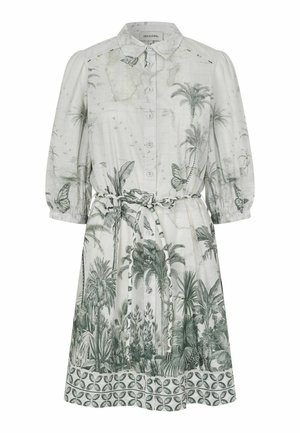 Jeune femme debout, mains derrière le dos, portant une robe blanche à imprimé botanique vert et des sandales marron sur un fond gris uni.