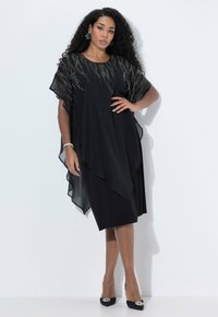 Femme aux longs cheveux bouclés portant une robe noire à plusieurs couches avec des détails en perles argentées et des talons noirs ornés de strass, se tenant contre un mur blanc.