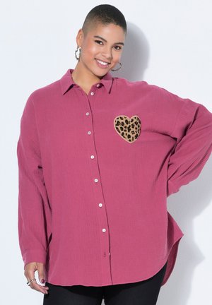 Smilende kvinde iført oversize magenta skjorte med leopardprint hjertepatch, øreringe i hoops og sorte bukser, poserer med hånden på hoften.
