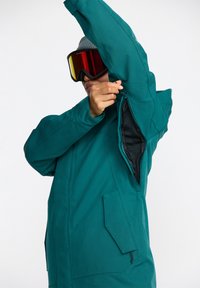 Veste de ski teal avec des zips aux manches, une doublure intérieure texturée et une coupe ajustée. Elle comporte des poches latérales et une capuche.