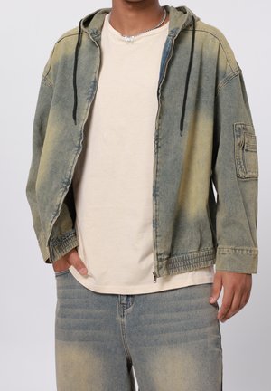 Osoba ubrana w wyblakłą zieloną kurtkę z zamkiem i kapturem, beżowy t-shirt, dopasowane wyblakłe dżinsy oraz srebrny łańcuszek z motylkiem jako zawieszką.