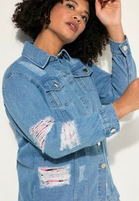 Giacca di jeans light blue con aree strappate e accenti rosa, bottoni argentati, colletto classico e due tasche sul petto.