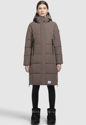 Př puffa bunda, �šedohnědá, po kolena, prošívaný design, vysoký límec s nastavovači, dvě zipové boční kapsy, nošena s černými legínami a botami.