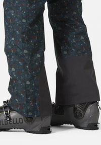 Waterdichte skibroek in donkerblauw met een gespikkeld patroon, met een grijze onderkant en zwarte skischoenen met verstelbare gespen.