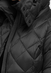 Chaqueta acolchada negra tipo puffer con cuello alto, que cuenta con cierre de cremallera y botones a presión, hecha de un material brillante y suave.