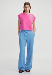 Roze cropped t-shirt met cap mouwen en bloemenversiering, gecombineerd met lichtblauwe wijde broek en zwarte sandalen.