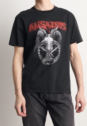 T-shirt in cotone nero con una grande grafica di un teschio di capra e corna, con testo rosso sopra, su uno sfondo grigio.