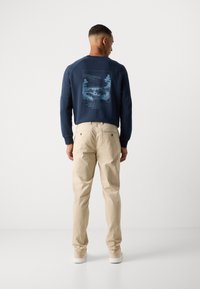 Man facing away wearing navy sweatshirt with blue nature scene and text, beige pants, and white sneakers on plain white background.