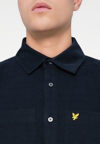 Tamno plava košulja s dugim rukavima s teksturiranim uzorkom, koja ima klasičan ovratnik i mali žuti izvezeni logo na džepu na prsima.