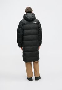 Crna zimska jakna s kapuljačom, s prošivenim uzorkom i logotipom The North Face na leđima. Nošena preko bež hlača i crnih cipela.