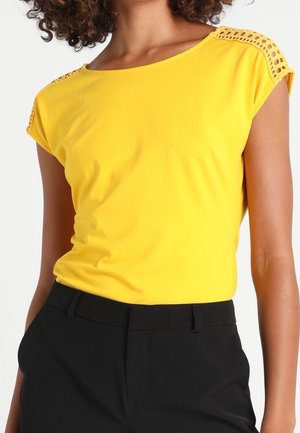 Femme portant un haut jaune à manches courtes avec des détails en dentelle sur les épaules et un pantalon noir taille haute.