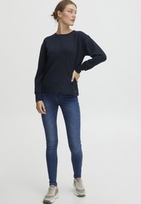 Mørkeblå sweatshirt med lange ermer og rund hals, kombinert med tettsittende blå jeans og nøytrale treningssko.