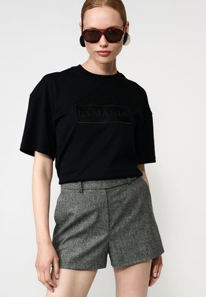 Juodas trumpas rankovėmis marškinėliai su reljefiniu "LAMANIA" logotipu, derinami su pilkais aukštu juosmeniu šortais tekstūruoto audinio. Rudi akiniai nuo saulės ir didelės auskarai.