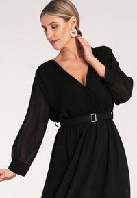 Czarna sukienka z plisowanym bodicem, długimi przezroczystymi rękawami i paskiem z kwadratową srebrną klamrą. Gładka faktura i zwiewny krój.