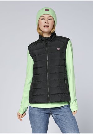 Schwarze Steppweste mit einem Reißverschluss vorn, die horizontale Steppabschnitte aufweist, kombiniert mit einem hellgrünen Langarmshirt und einer weichen grünen Strickmütze.