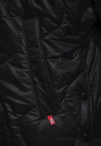 Czarna pikowana kurtka o gładkiej fakturze, z wyraźnymi wzorami przeszyć, przednią kieszenią i widoczną czerwoną metką marki. W komplecie czarne guziki.