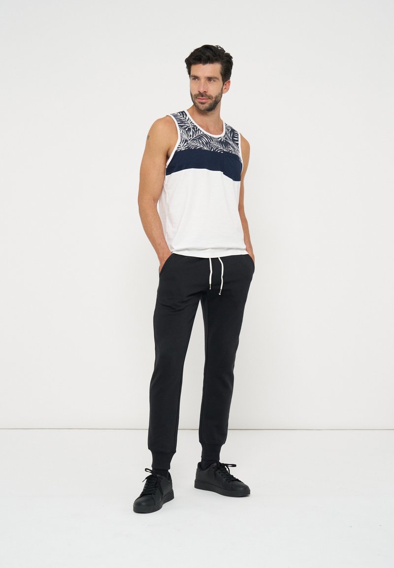 Homme debout les mains dans les poches portant un débardeur blanc et bleu marine à motifs, un pantalon jogger noir et des baskets noires.