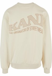 Kremgenser med ribbestrikkede mansjetter og nedkant, med et stort "KANI"-grafikk i en teksturert design og teksten "THE ORIGINATOR OF STREETWEAR".