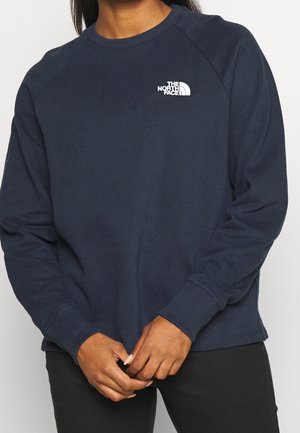 Person trägt einen marineblauen Sweatshirt mit Rundhalsausschnitt und dem The North Face Logo auf der Brust, steht mit vor der Brust verschränkten Händen.