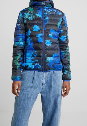 Person iført en sort dynejakke med blå og grønne blomsterprint og en lynlås foran, kombineret med løse lyseblå denimjeans.