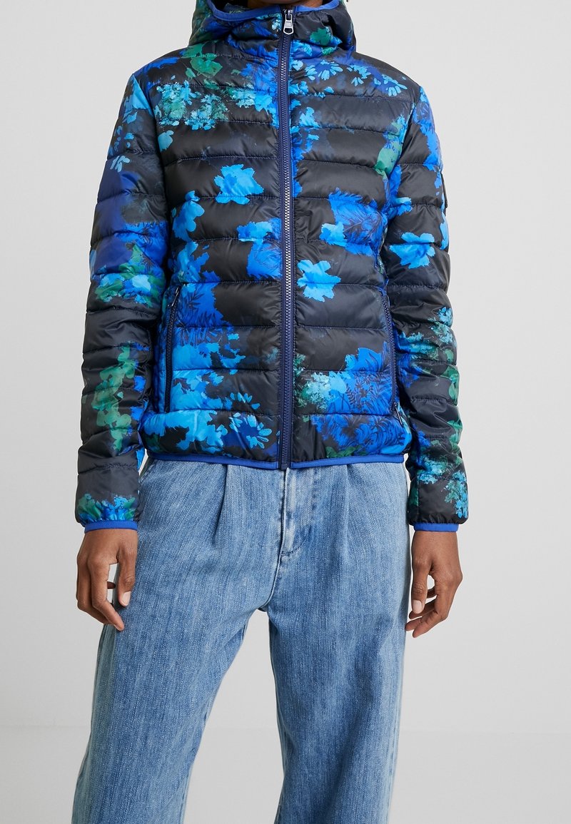 Personne portant une doudoune noire avec des motifs floraux bleus et verts et une fermeture éclair devant, associée à un jean en denim bleu clair et ample.