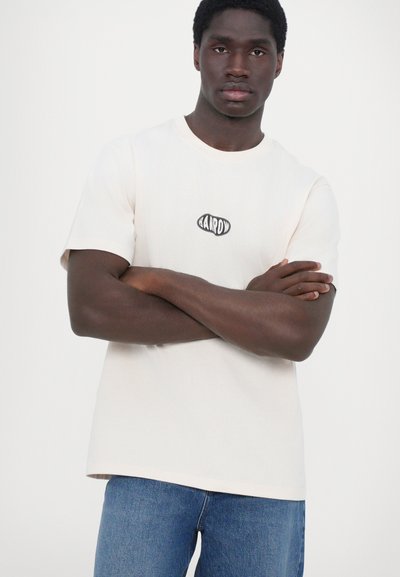 T-shirt en coton rose clair à manches courtes, avec un logo ovale noir sur la poitrine. Associé à un jean en denim bleu.