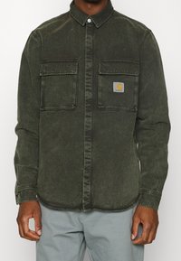 Ciemnozielona koszula dżinsowa z kołnierzykiem zapinanym na guziki, dwiema kieszeniami na piersi oraz małym logo Carhartt na lewej kieszeni. Gładka struktura.