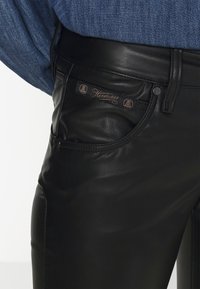 Czarne skórzane spodnie o gładkiej fakturze, z przednią kieszenią i metką z logo. Pas ma klasyczne zapięcie na guzik.