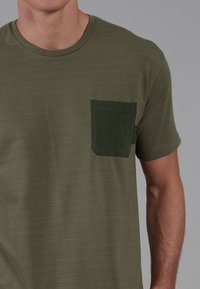 Camiseta de manga corta color verde oliva con un bolsillo en el pecho de un verde oscuro texturizado y un escote redondo, elaborada en una tela ligera.