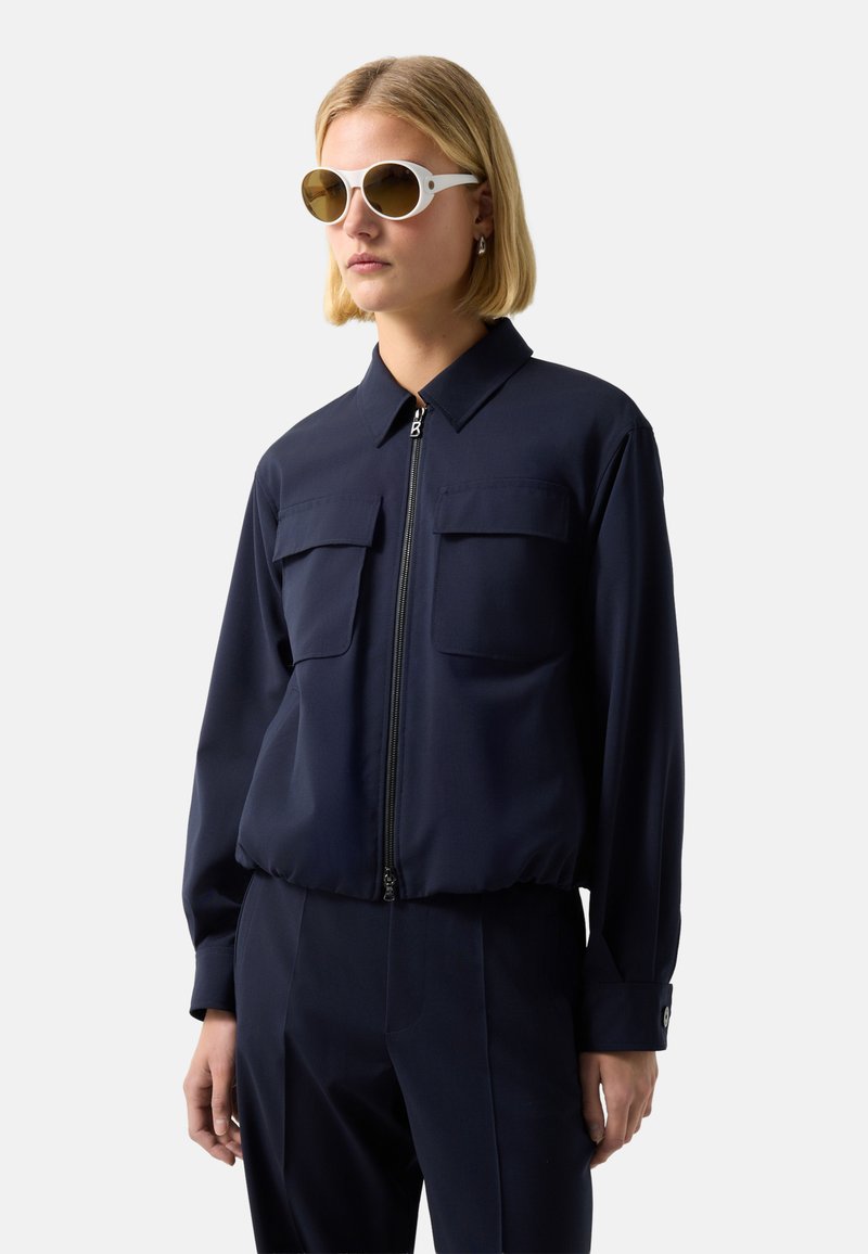 Blonde woman wearing white sunglasses and a navy zip-up jacket with flap pockets, standing against a white background.