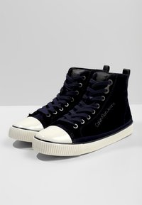 Korkeat mustat samettikengät, joissa on valkoiset kumi kärjet ja pohjat, mustat nauhat sekä "Calvin Klein Jeans" -logo sivussa.